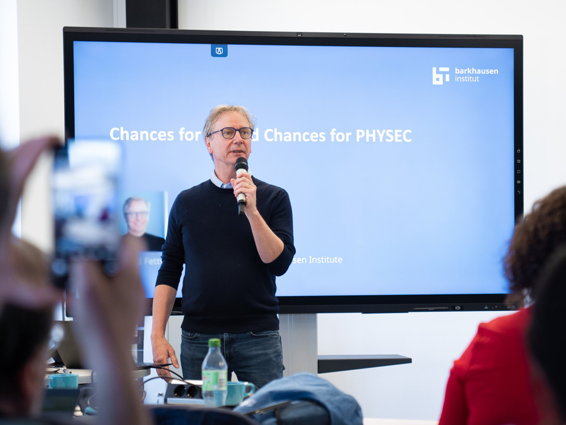 Prof. Gerhard Fettweis steht vor einem Bildschirm. darauf ist eine Präsentation zu sehen mit weißer Schrift auf blauem Hintergrund. Prof. Fettweis hält ein Mikrofon in der Hand. Im Vordergrund ist unscharf zu sehen: zwei Hände, die ein Smartphone auf Gerhard gerichtet halten. Rechts sind 2 Personen leicht angeschnitten mit ihrem Rücken/Hinterkopf zu sehen.