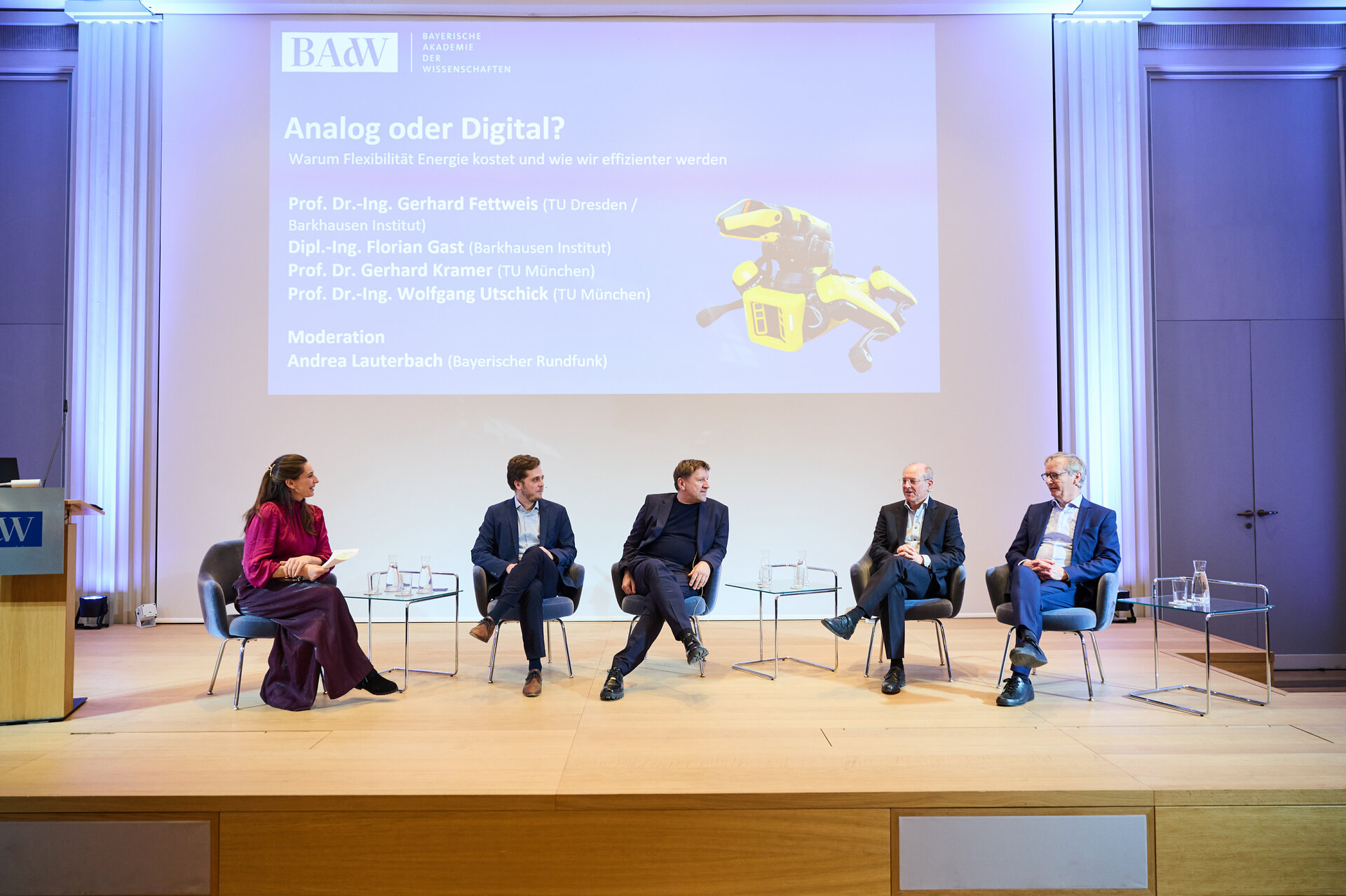 Five people seated on a stage in a panel discussion setup with a large screen behind them showing a presentation slide titled 'Analog oder Digital'