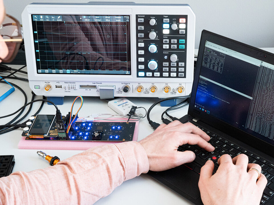 Chip Masur25 wird auf einem Evaluationsboard getestet. Im Vordergrund tippt eine Person auf einer Tastatur eines Laptops, der mehrere Konsolenfenster mit Code oder Messdaten anzeigt. Das Evaluationsboard ist mit mehreren Kabeln und Bauteilen bestückt und zeigt leuchtende blaue LEDs. Im Hintergrund steht ein Oszilloskop mit einem großen Display, auf dem mehrere Kurven und Messwerte sichtbar sind. Die Szene vermittelt eine typische Laborsituation für Elektroniktests.
