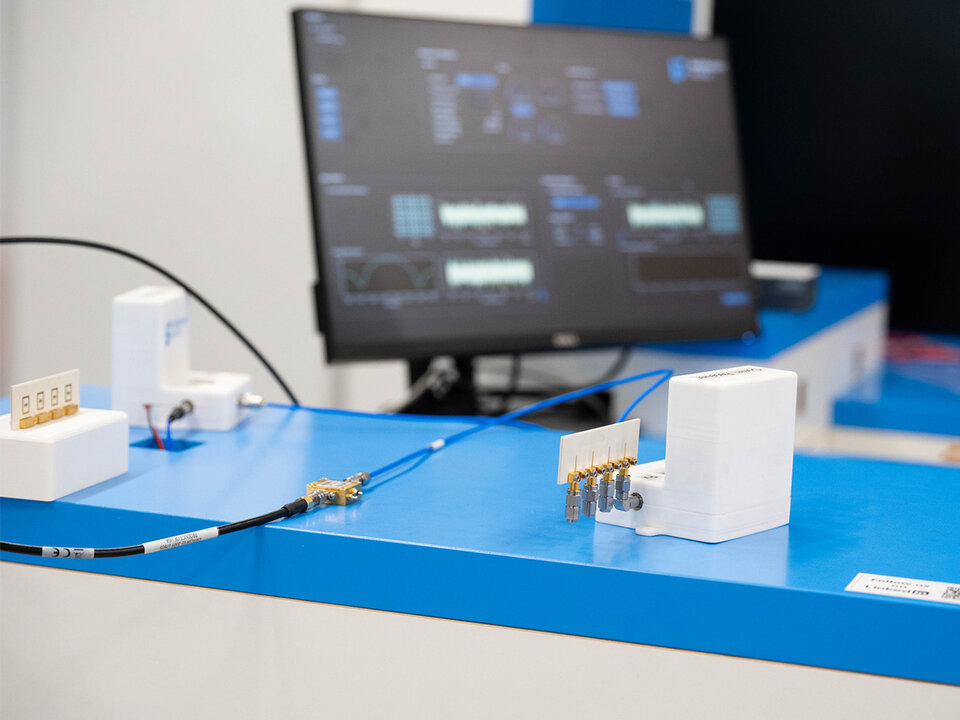 Electronic testing setup with white connectors and cables on a blue table and a monitor displaying software interface in the background