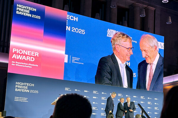 The three men stand on the stage for a group photo. The large screen behind the stage shows Prof. Schwaiger's congratulations.