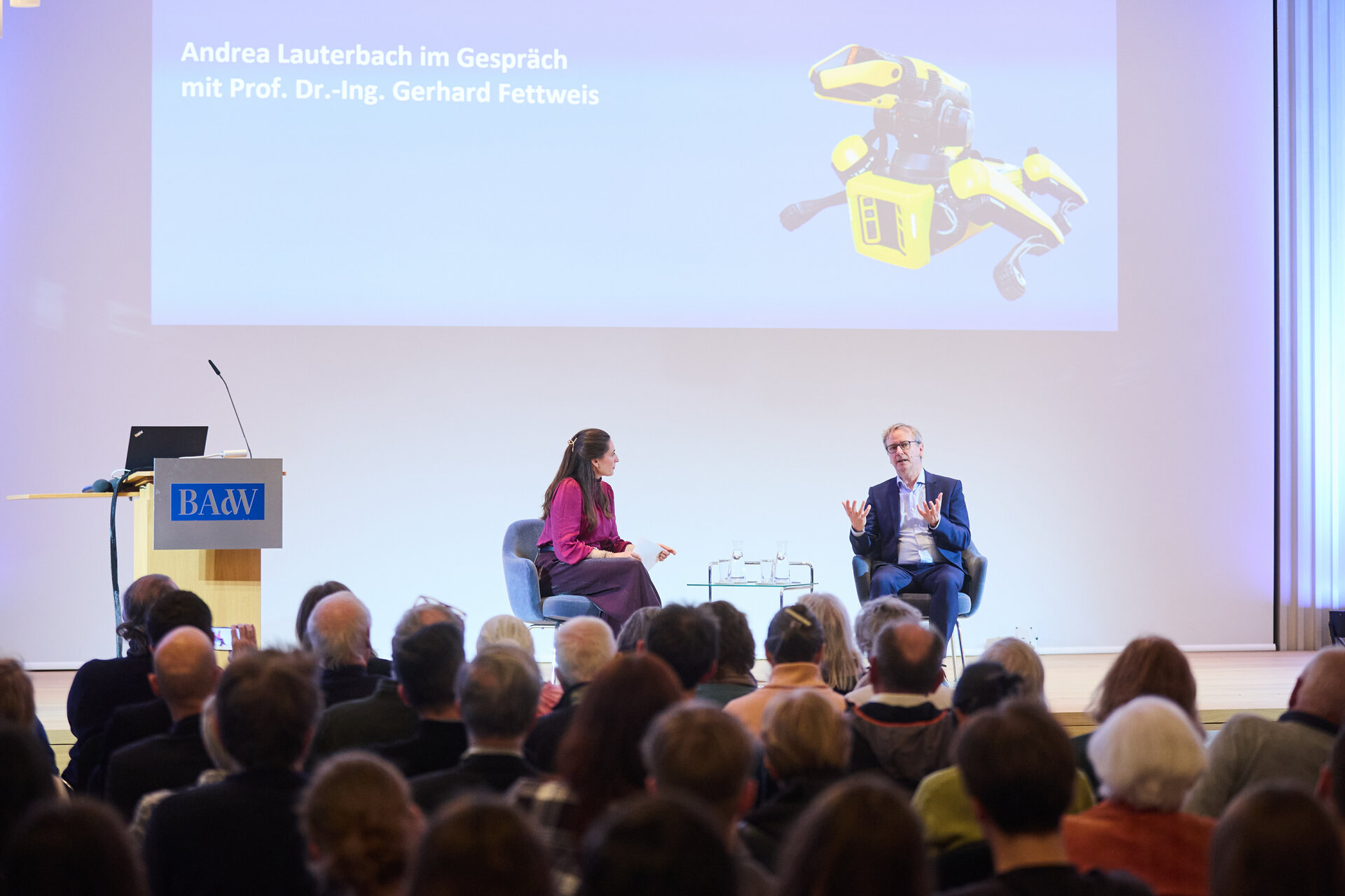 Two people seated on stage in front of an audience with a screen behind them showing a yellow robotic dog and text in German