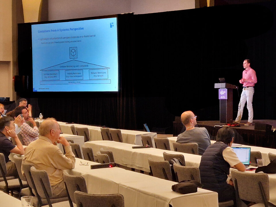 Till Miemietz präsentiert auf der Bühne bei USENIX25 eine Folie zum Thema Container aus Systemperspektive vor Publikum in einem Konferenzraum.