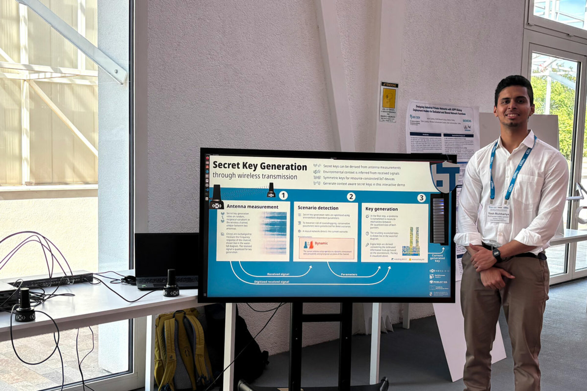 Ein Mann in einem weißen Hemd, mit einem Lanyard um den Hals und einem Namensschild mit der Aufschrift 'Yash Richhariya', steht neben einem großen Monitor, auf dem eine Präsentation mit dem Titel 'Secret Key Generation through wireless transmission' mit Diagrammen und Text angezeigt wird.