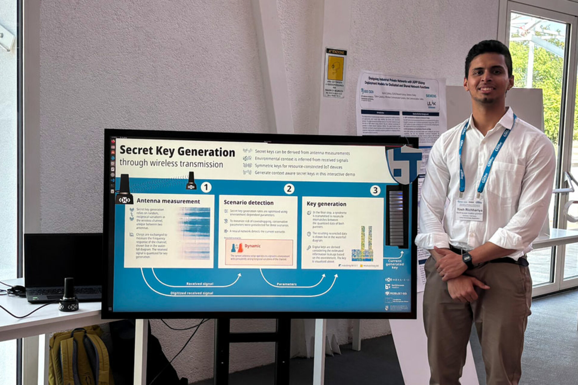 A Man dressed in a white Shirt, with a Lanyard around his neck, holding a name badge with the name 'Yash Richhariya' stands next to a large monitor displaying a presentation titled 'Secret Key Generation through wireless transmission' with diagrams and text.