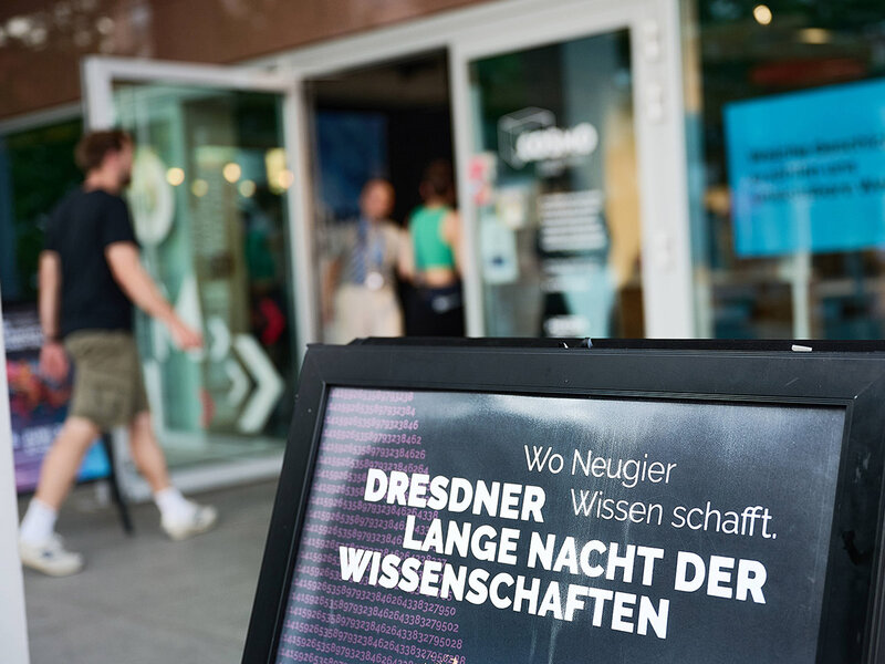 Aufsteller mit dem Text 'Wo Neugier Wissen schafft. Dresdner Lange Nacht der Wissenschaften' vor einem Gebäude mit geöffneten Glastüren und mehreren Personen im Hintergrund.