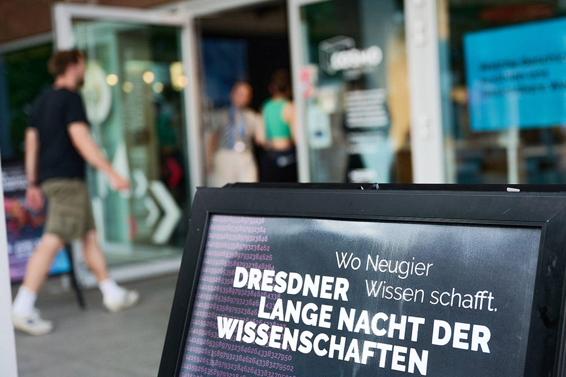 Aufsteller mit dem Text 'Wo Neugier Wissen schafft. Dresdner Lange Nacht der Wissenschaften' vor einem Gebäude mit geöffneten Glastüren und mehreren Personen im Hintergrund.