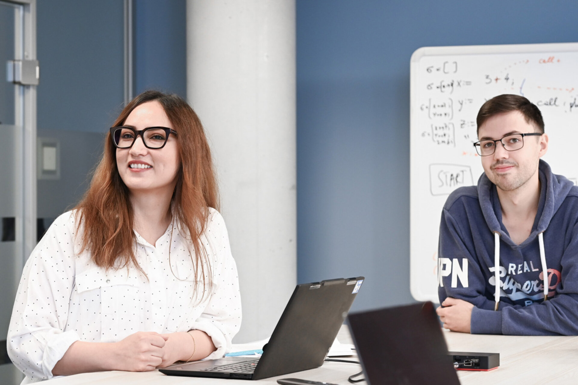 Zwei Personen sitzen an einem Tisch mit geöffneten Laptops, im Hintergrund eine Tafel mit mathematischen Formeln.