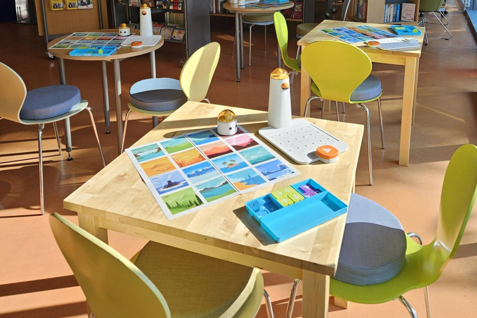 Mehrere Tische mit gelben Stühlen in einem hellen Raum, auf einem Tisch liegen bunte Karten und kleine Roboterbauteile für einen Robo Kids Workshop