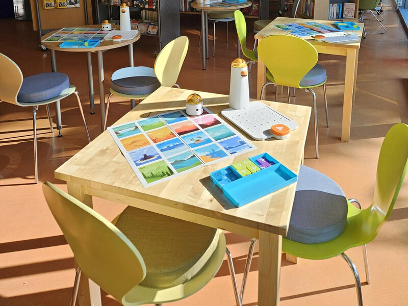 Mehrere Tische mit gelben Stühlen in einem hellen Raum, auf einem Tisch liegen bunte Karten und kleine Roboterbauteile für einen Robo Kids Workshop