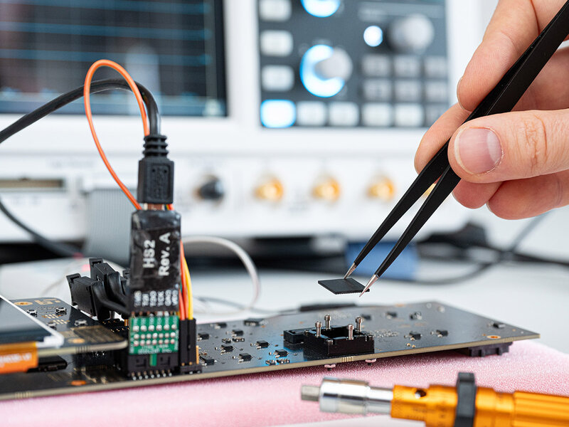Eine Hand hält mit einer Pinzette einen Chip, der gerade in den Sockel eines Evaluationsboards eingesetzt wird. Das Evaluationsboard liegt auf einer rosafarbenen Unterlage. Mehrere Kabel sind mit dem Board verbunden, darunter ein orangefarbenes und ein schwarzes Kabel. Im Hintergrund sind unscharf ein Oszilloskop und weitere elektronische Messgeräte zu erkennen.