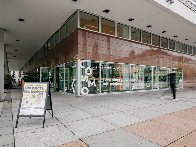 Eingang des COSMO Wissenschaftsforums mit Glasfassade und Aufsteller zur Ausstellung 'Mensch, Roboter!' auf dem Gehweg.
