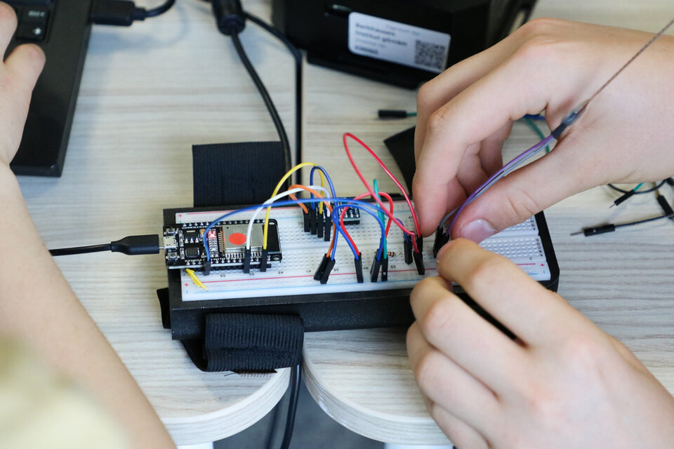 Hands connecting colored wires to a breadboard with electronic components on a table. A laptop is visible, suggesting a tech project.