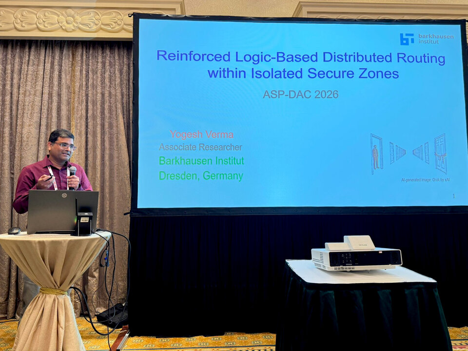 Man standing at a podium with a laptop giving a presentation titled 'Reinforced Logic-Based Distributed Routing within Isolated Secure Zones' with text including 'Yogesh Verma, Associate Researcher, Barkhausen Institut, Dresden, Germany' and 'ASP-DAC 2026'