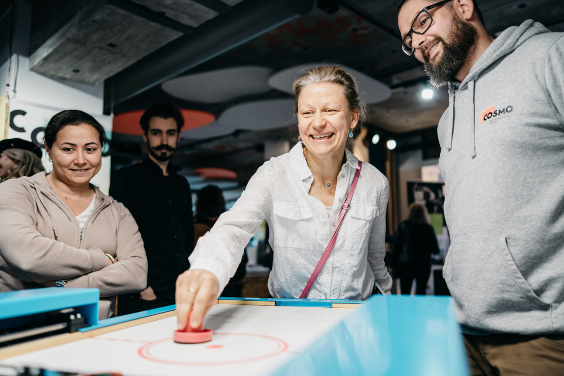 Person in weißem Hemd spielt an einem Airhockey-Tisch, umgeben von weiteren Personen in einem Ausstellungsraum.