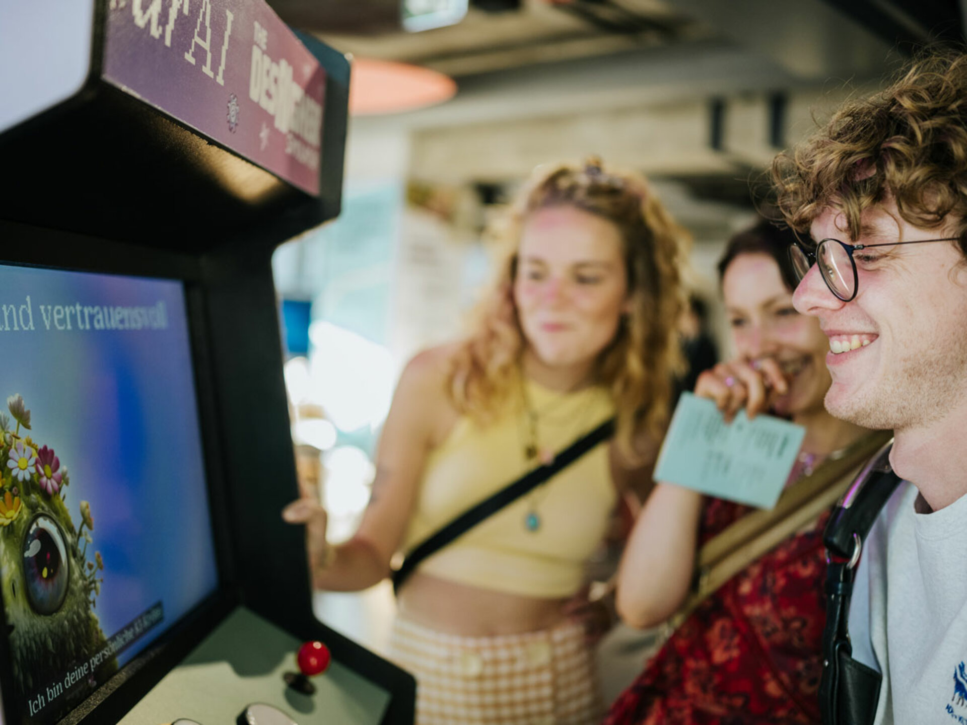 Eine junger Mann bedient einen Spieleautomaten zum Thema Künstlicher Intelligienz während zwei weitere Personen zusehen.