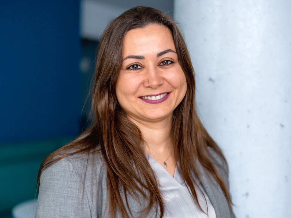 Prof. Elif Bilge Kavun trägt einen hellgrauen Blazer und ein helles Oberteil. Langes, braunes Haar fällt über die Schultern. Im Hintergrund sind eine weiße Säule und eine blaue Wand zu sehen.