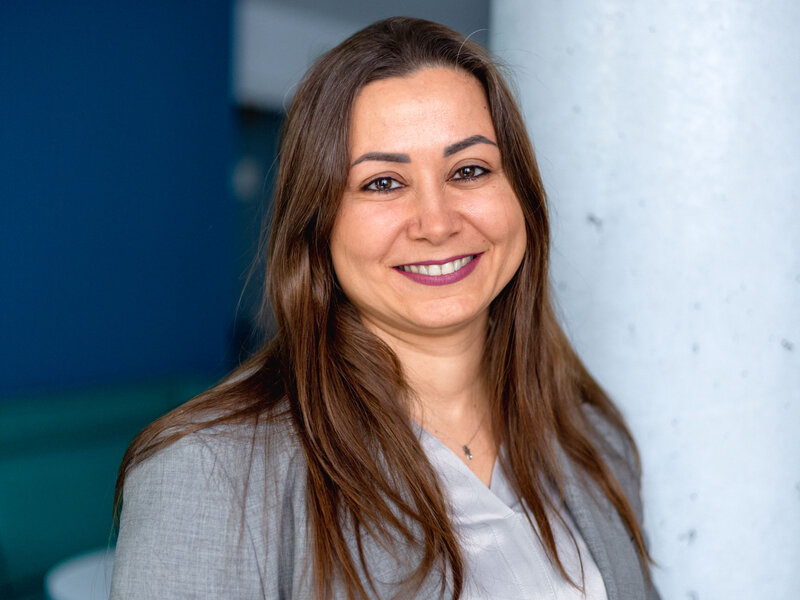 Prof. Elif Bilge Kavun trägt einen hellgrauen Blazer und ein helles Oberteil. Langes, braunes Haar fällt über die Schultern. Im Hintergrund sind eine weiße Säule und eine blaue Wand zu sehen.