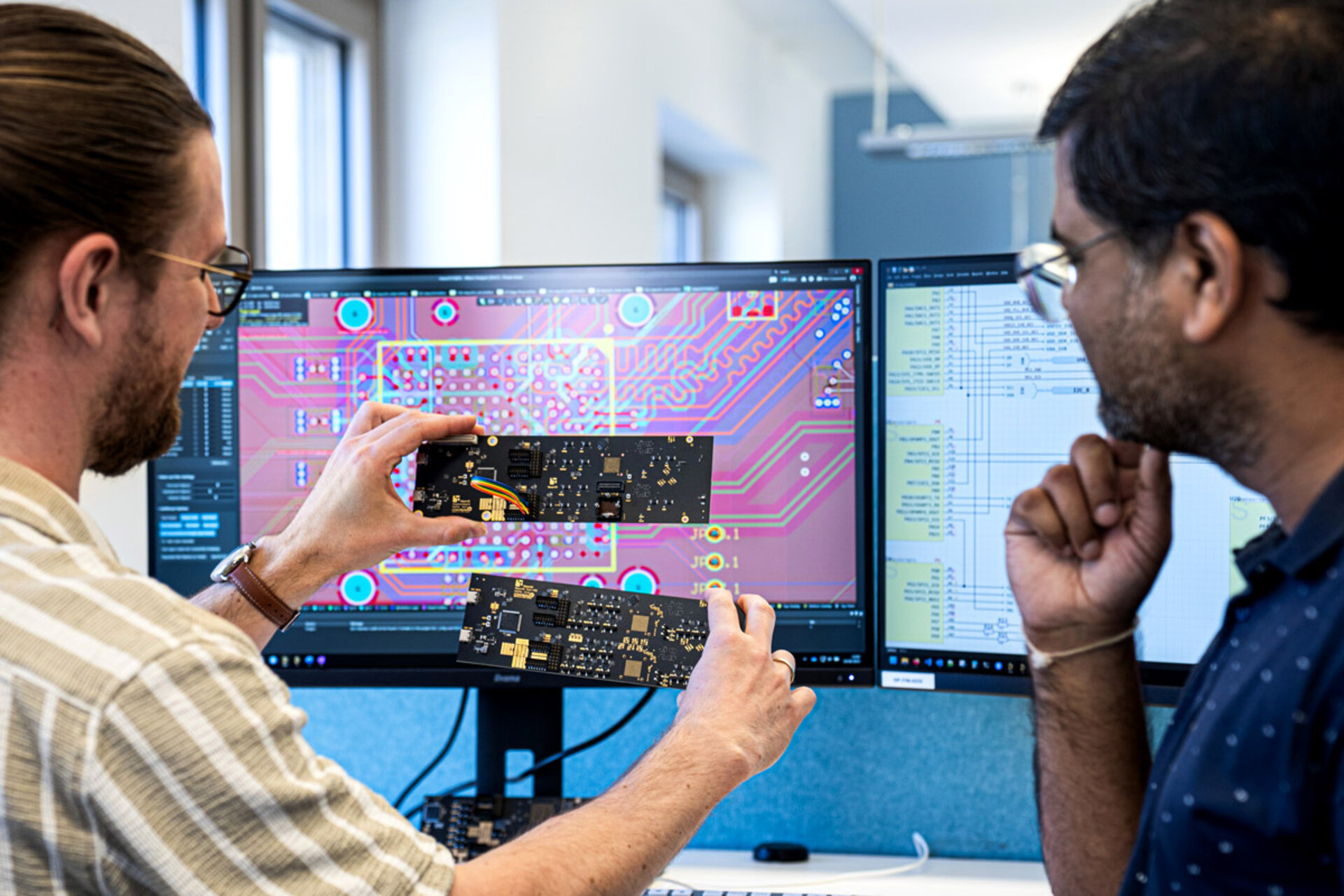 Zwei Personen vergleichen eine Leiterplatte vor einem Bildschirm mit Leiterplatten-Designsoftware in einem Büro.
