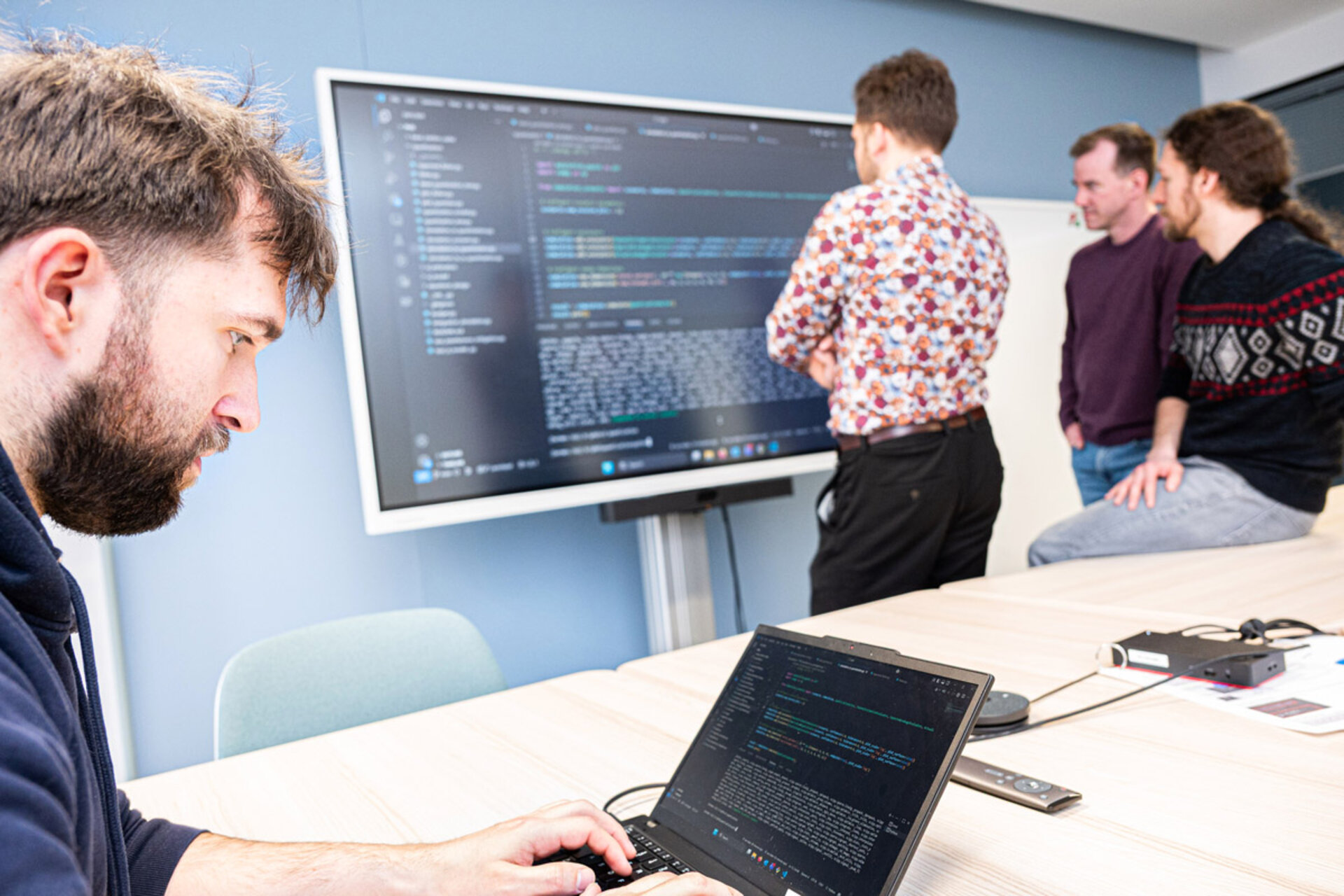 Person sitzt an einem Tisch und tippt auf einem Laptop, im Hintergrund drei Personen vor einem großen Bildschirm mit Programmiercode.