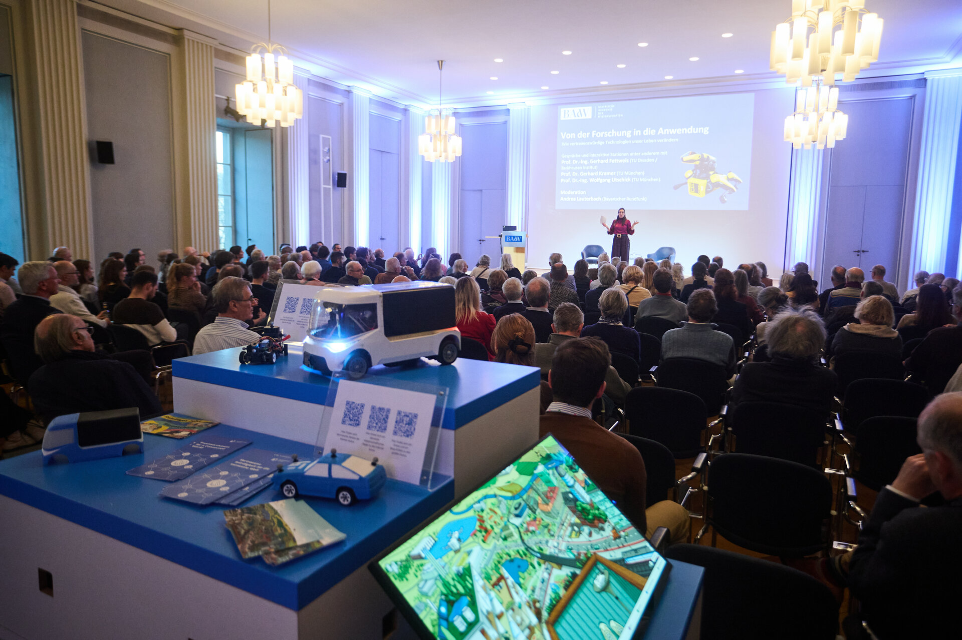 Speaker presenting to an audience in a conference room with a model vehicle and digital display in the foreground