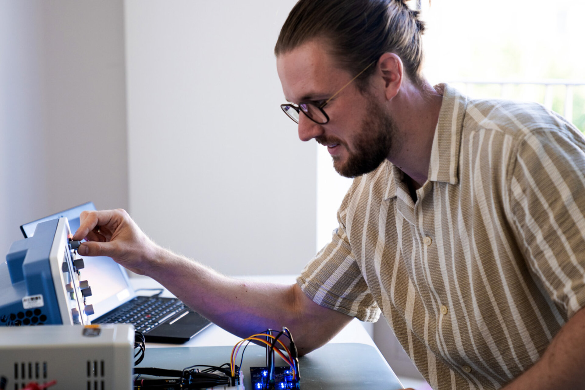 Person mit gestreiftem Hemd und Armbanduhr bedient ein elektronisches Messgerät an einem Tisch mit Schaltkreis und Laptop