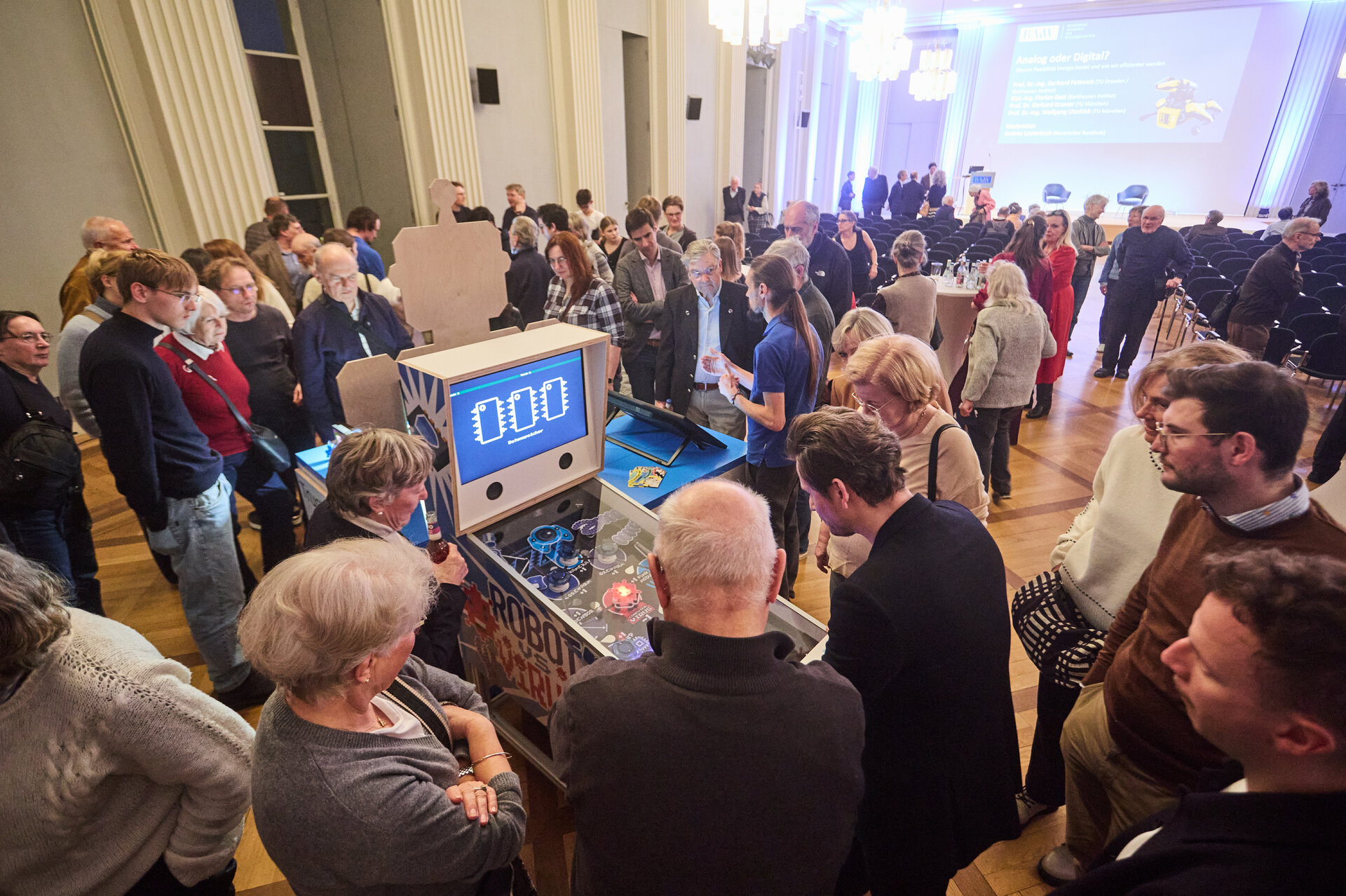 Group of people gathered around a table with a digital display showing '0.00' in a large room with chandeliers and a presentation screen in the background