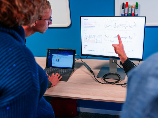 Zwei Personen betrachten die Anzeige auf einem großen Monitor, der technische Diagramme und Messdaten zeigt. Eine Person deutet auf die Visualisierung, während eine andere an einem Laptop arbeitet. Im Hintergrund ist eine blaue Wand mit einem Whiteboard zu erkennen.