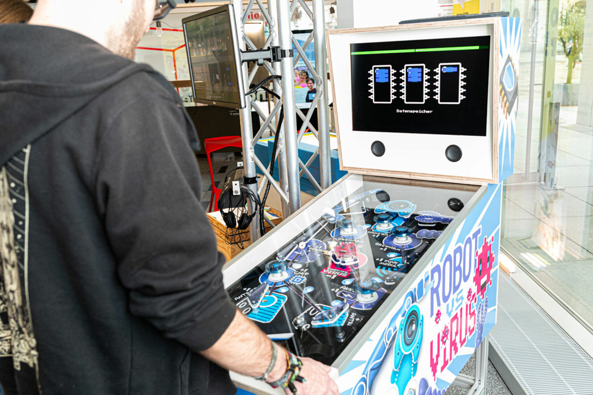 Person bedient einen blauen, mit Robotik-Motiven verzierten Flipperautomaten mit Bildschirm und zwei schwarzen Knöpfen im COSMO Wissenschaftsforum.