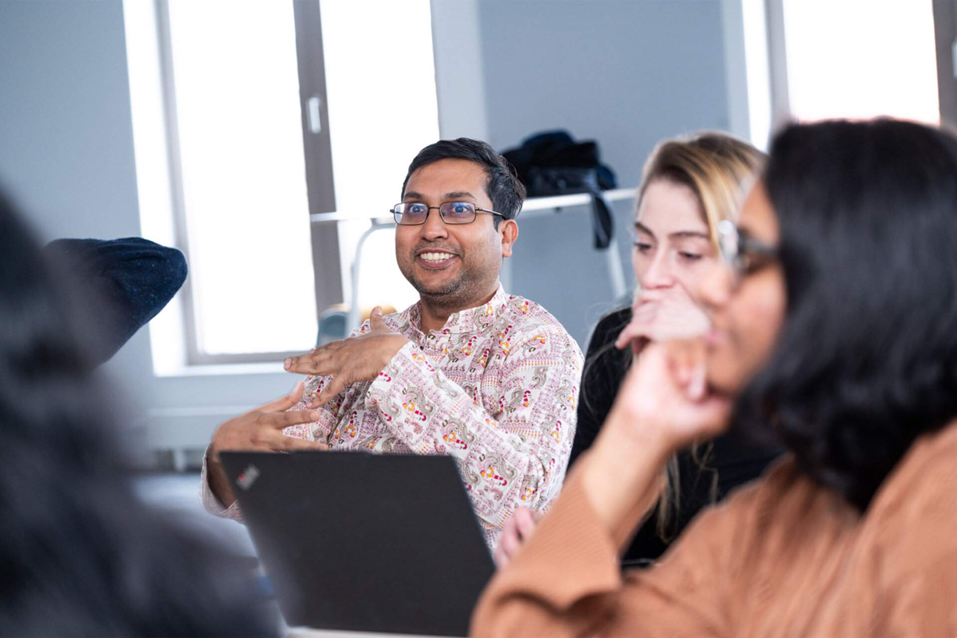 Man in patterned shirt gesturing while sitting at a table with a laptop in front of him and two blurred people nearby