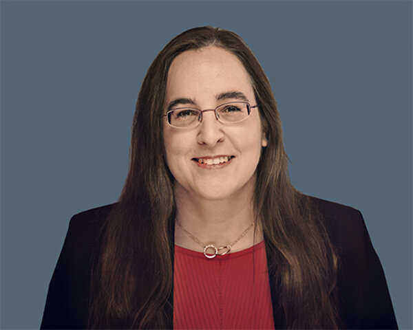 Frau mit langen braunen Haaren trägt eine schwarze Jacke, ein rotes Oberteil und eine Halskette mit zwei ineinander verschlungenen Ringen vor blau-grauem Hintergrund.