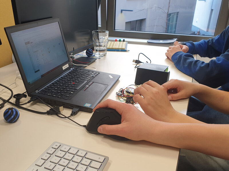 Hände, die eine schwarze Computermaus neben einem Laptop bedienen, auf dem eine Software-Oberfläche angezeigt wird, mit elektronischen Bauteilen und Kabeln auf dem Schreibtisch.