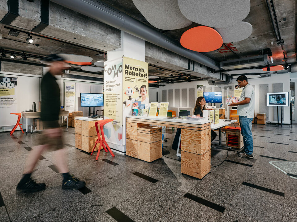 Innenraum des COSMO Wissenschaftsforums mit Ausstellung 'Mensch, Roboter!'. Tisch mit Informationsmaterial, zwei Personen, Deckenakustikelemente in Grau und Orange, sichtbare Technik und Betonwände.