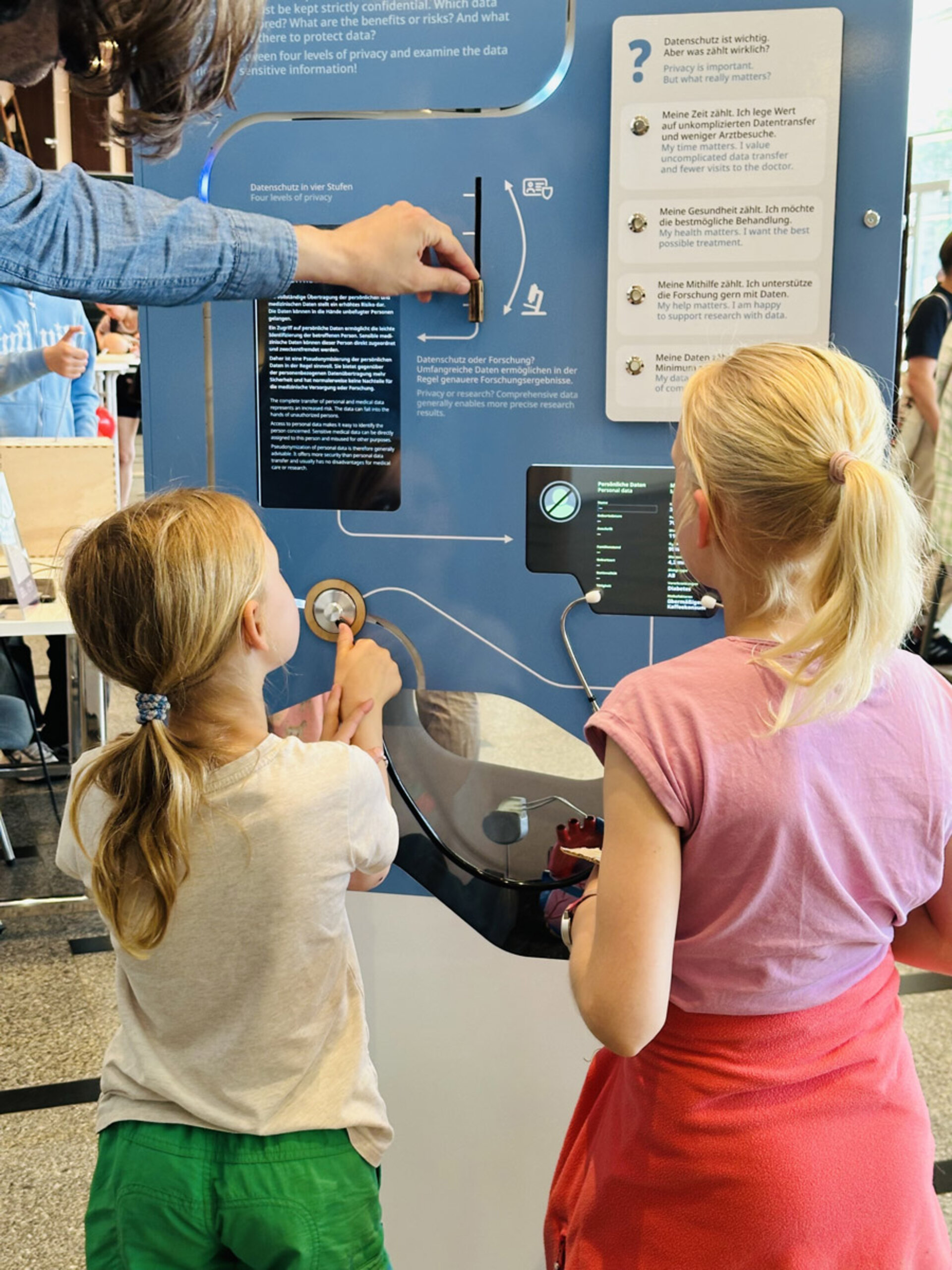 Zwei Kinder interagieren mit einer lehrreichen Ausstellung zum Thema Datenschutz, bei der der Schwerpunkt auf ansprechenden Displays und praktischen Elementen liegt. Die Hand eines Erwachsenen stellt einen Knopf ein und leitet die Kinder bei ihrer Erkundung in einer hellen, interaktiven Umgebung.