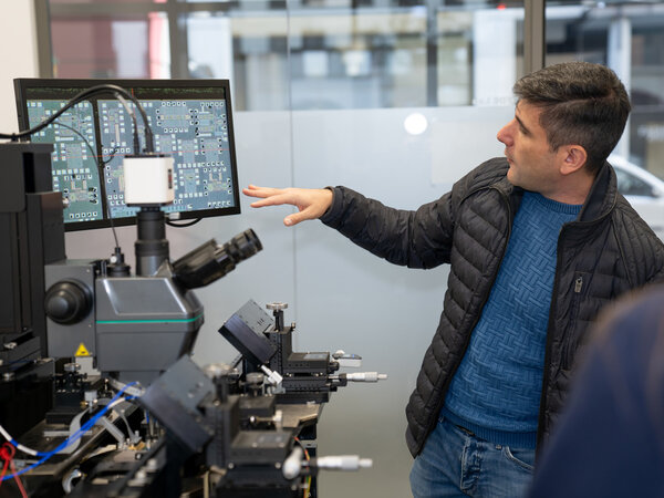 The picture shows a man standing in front of a complex microscope and pointing at the screen. The screen shows a detailed image of a microelectronic component. The man appears to be explaining the technology to a colleague. The image conveys an impression of modern technology and technical know-how.