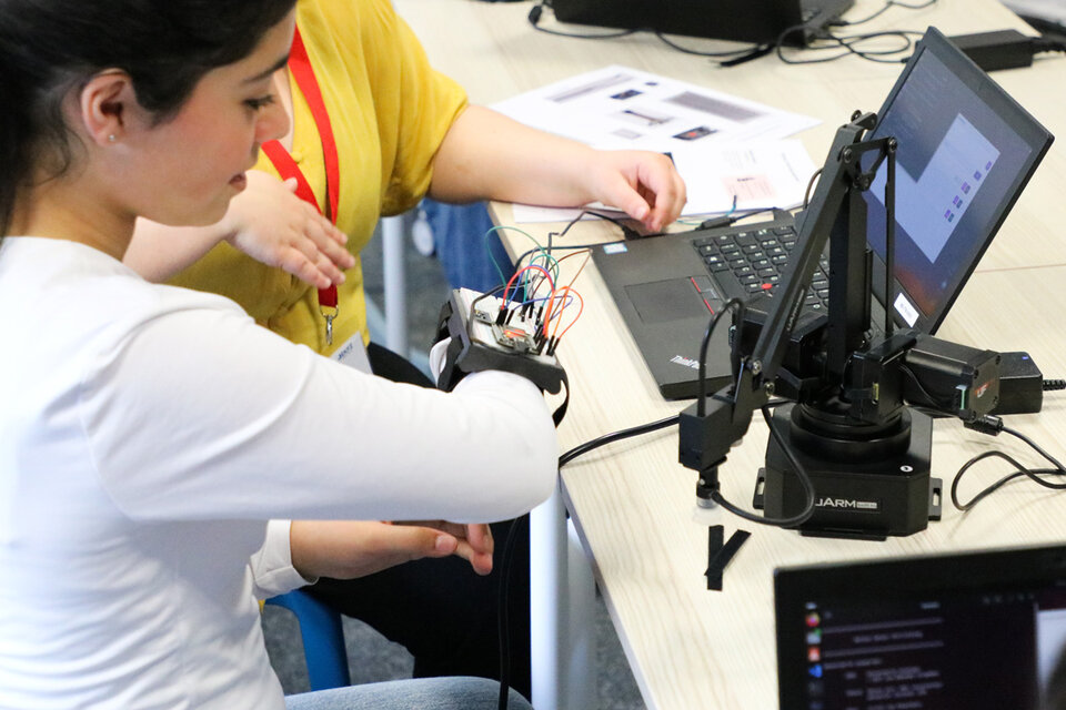 A woman in a white shirt works with electronic components attached to her wrist, connected to a laptop on a desk, collaborating with another person.