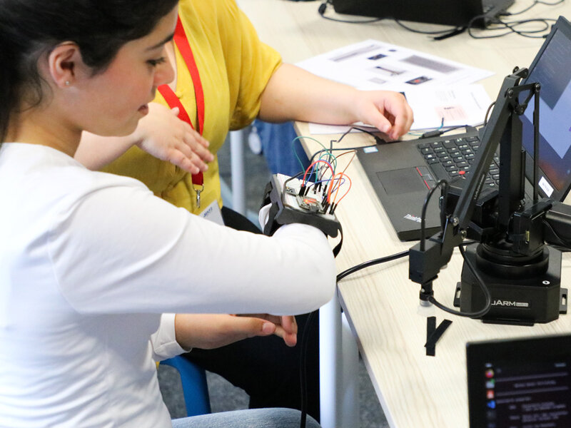 A woman in a white shirt works with electronic components attached to her wrist, connected to a laptop on a desk, collaborating with another person.