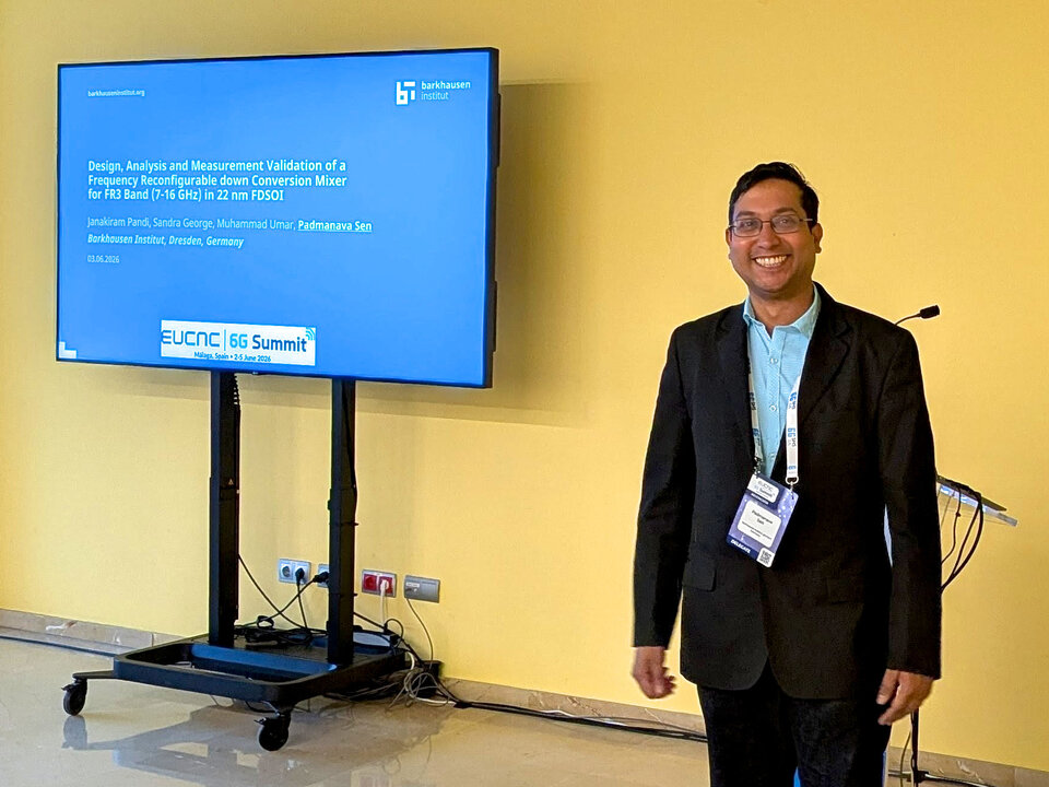 Man in a suit and conference badge standing next to a large screen displaying a technical presentation slide in a yellow room