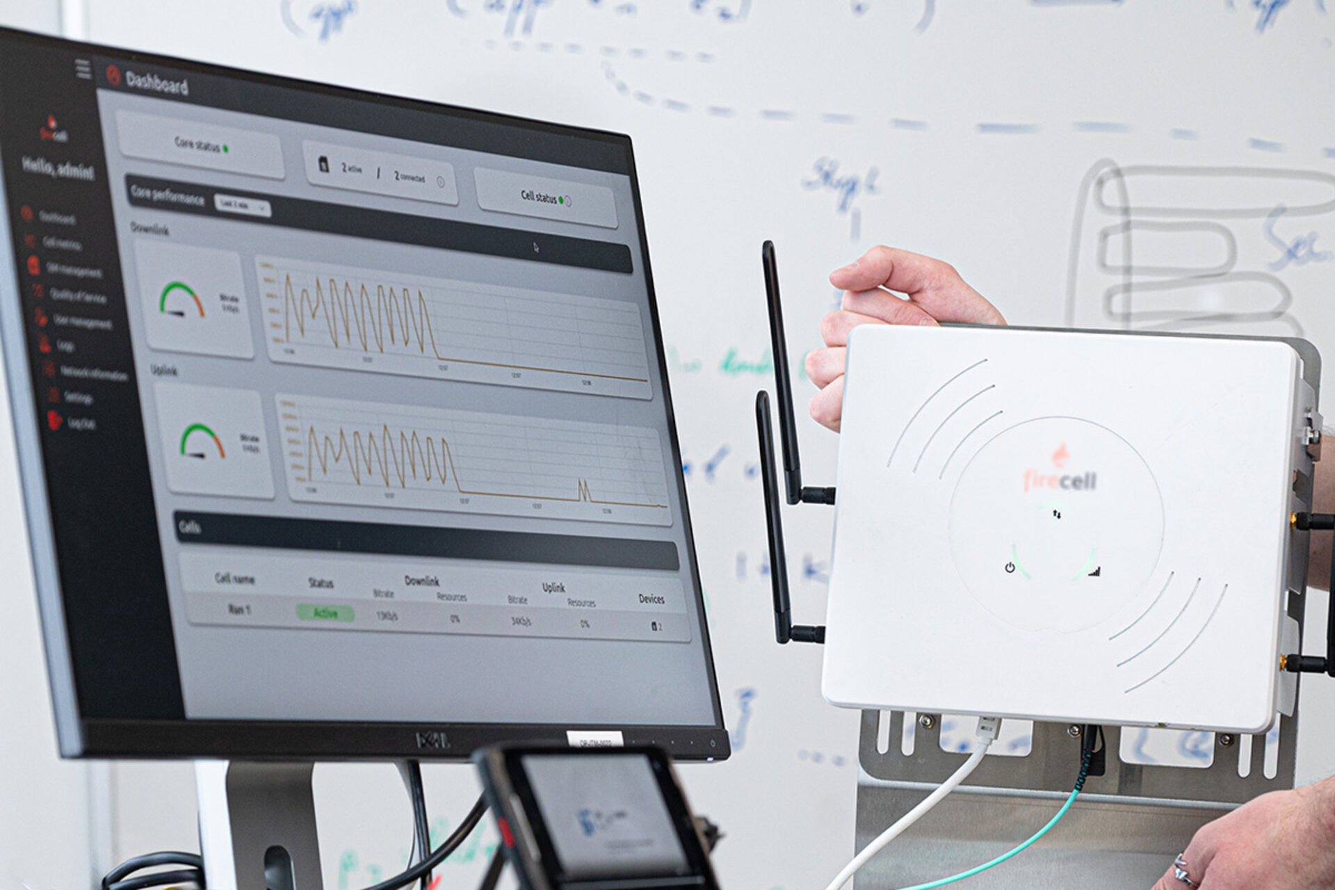 Hand hält ein weißes Gerät mit Antenne vor einen Monitor mit Dashboard-Anzeige und Grafiken im Hintergrund