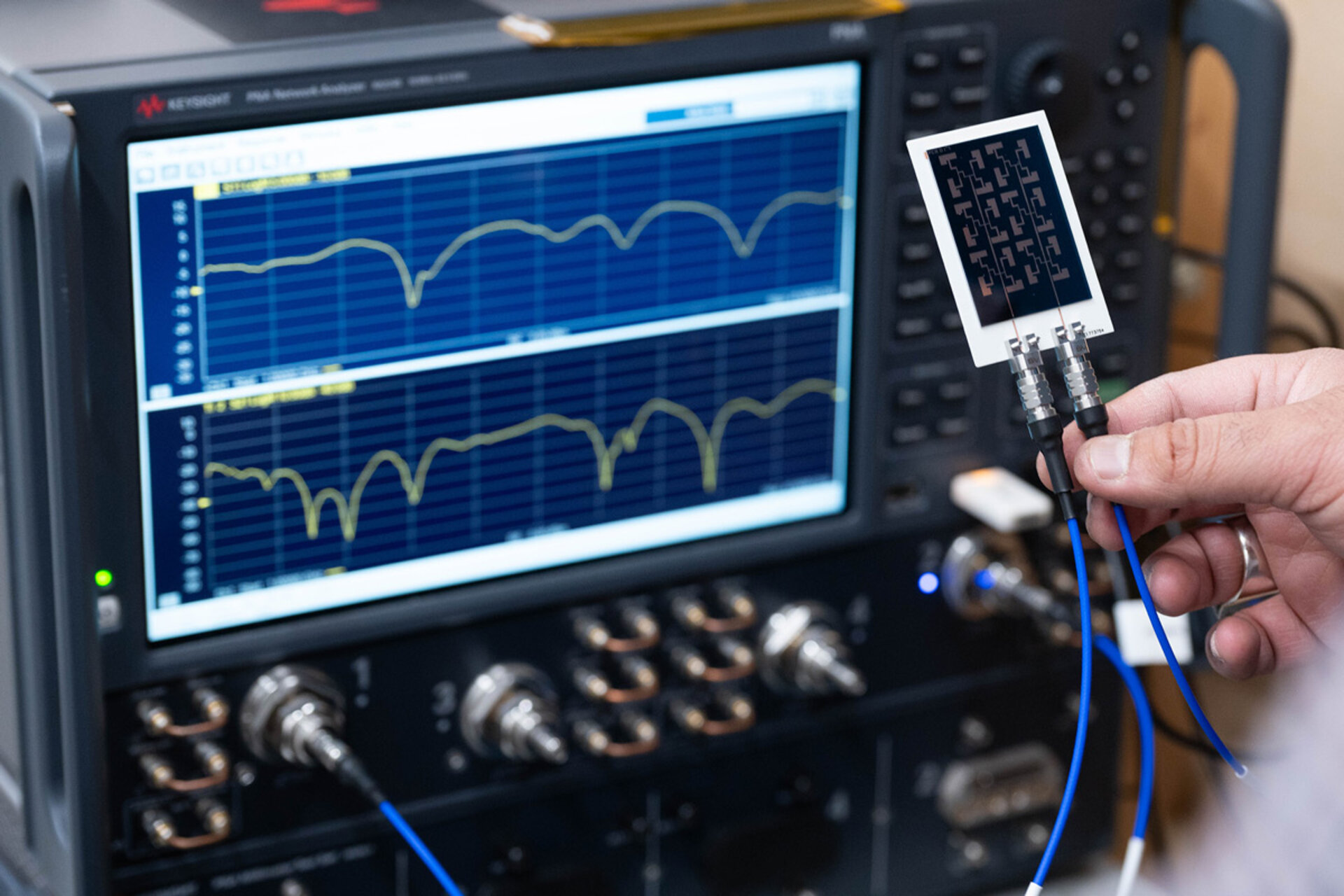 Hand holding a small circuit board with connectors in front of a screen displaying waveforms and electronic measurement equipment