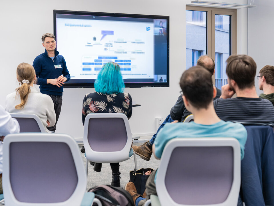 One person is standing in front if a huge monitor explaining something. In the foreground you can see people sitting on chairs listening to that person.