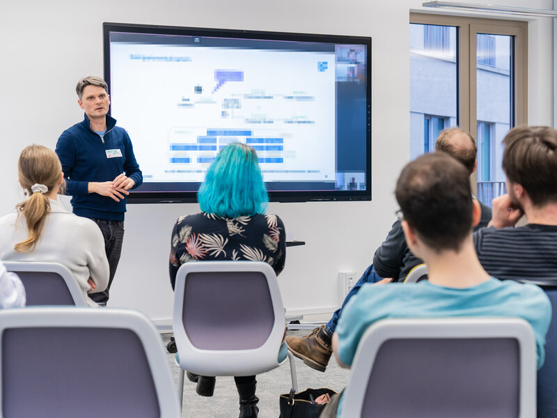 One person is standing in front if a huge monitor explaining something. In the foreground you can see people sitting on chairs listening to that person.
