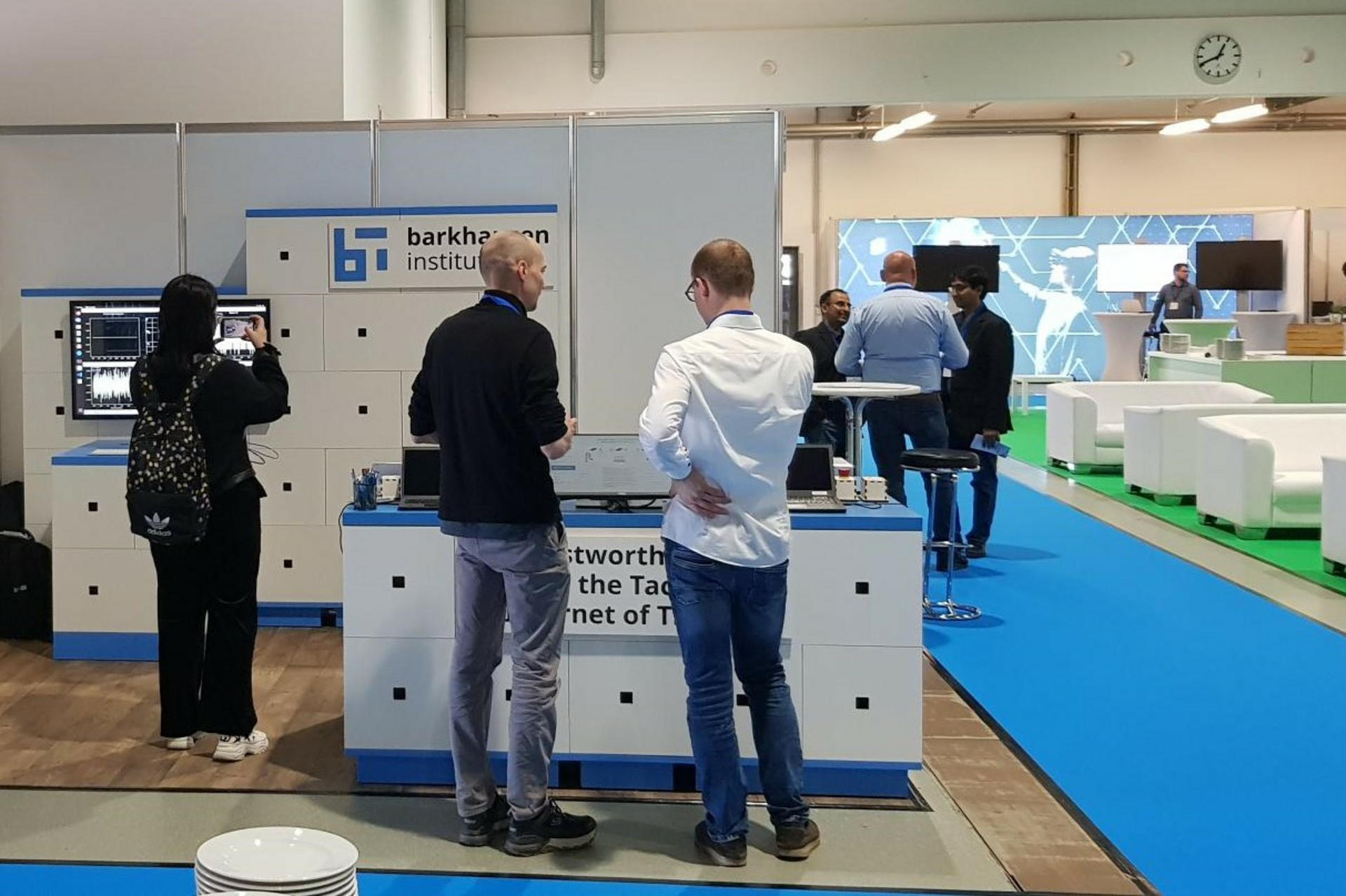 3 Leute stehen am Stand des Barkhausen Institutes. Eine Person macht ein Foto der Demo. Zwei andere unterhalten sich.