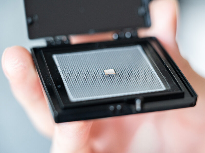 Hand hält eine geöffnete schwarze Kunststoffbox mit einem kleinen, quadratischen Chip ohne Gehäuse auf einer perforierten Trägerplatte