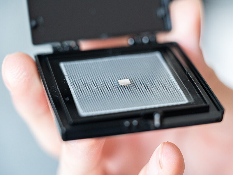 Hand holding an open black plastic case containing a small square bare chip on a perforated carrier plate