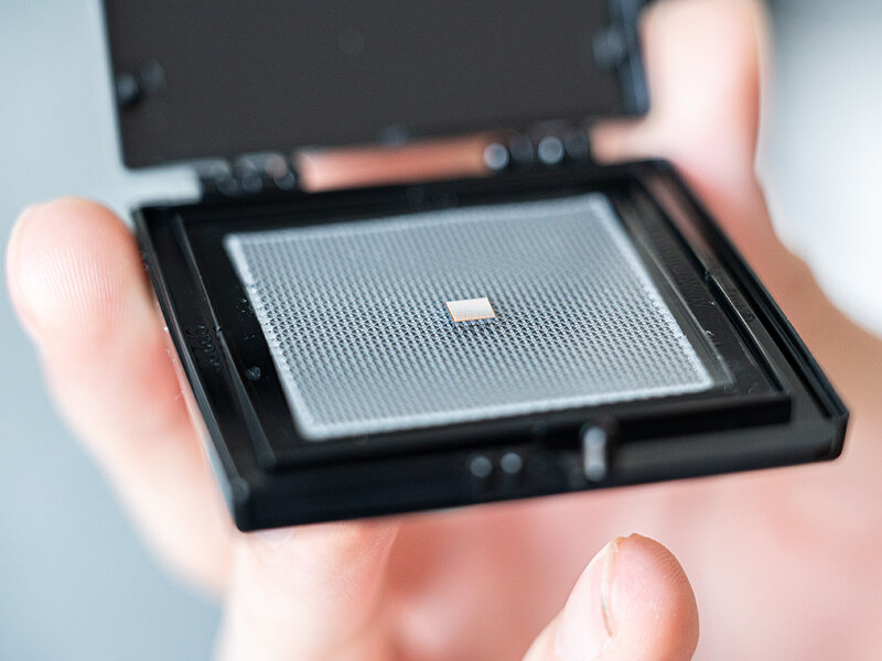 Hand hält eine geöffnete schwarze Kunststoffbox mit einem kleinen, quadratischen Chip ohne Gehäuse auf einer perforierten Trägerplatte