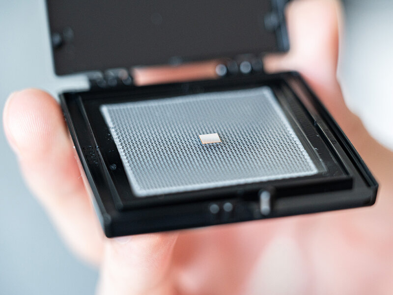 Hand holding an open black plastic case containing a small square bare chip on a perforated carrier plate