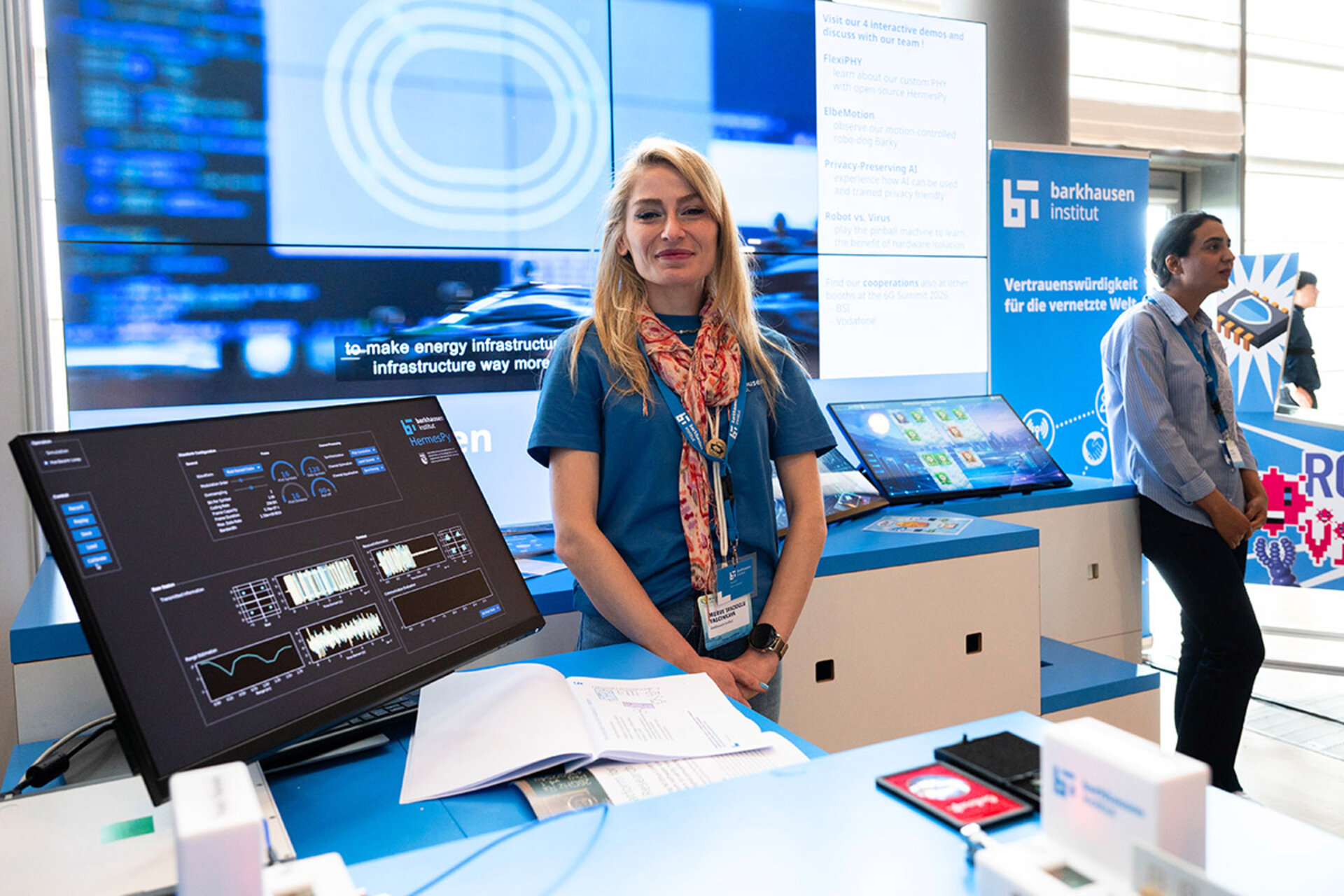 Woman standing behind a table with multiple digital screens and open documents at a technology exhibition booth