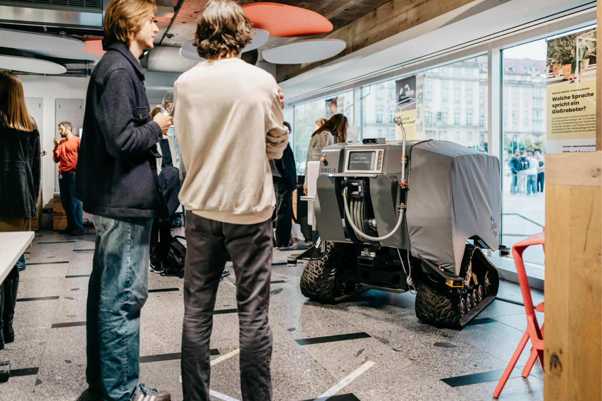 Two men stand in front of a small robot with a gray cover and tracked wheels at an exhibition in the COSMO Science Forum.