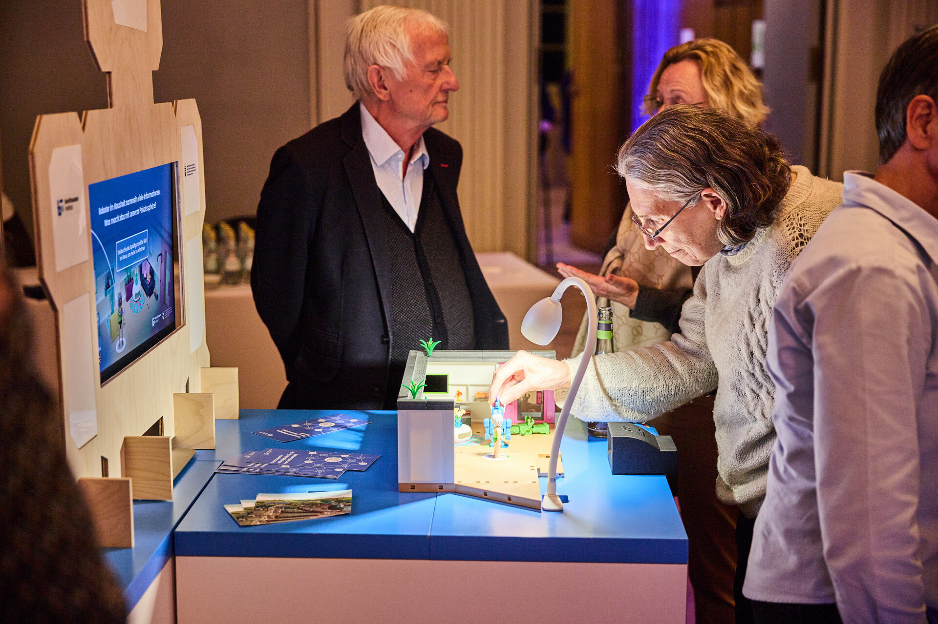 Older man and woman interacting with a tabletop display in a dimly lit room with other people nearby